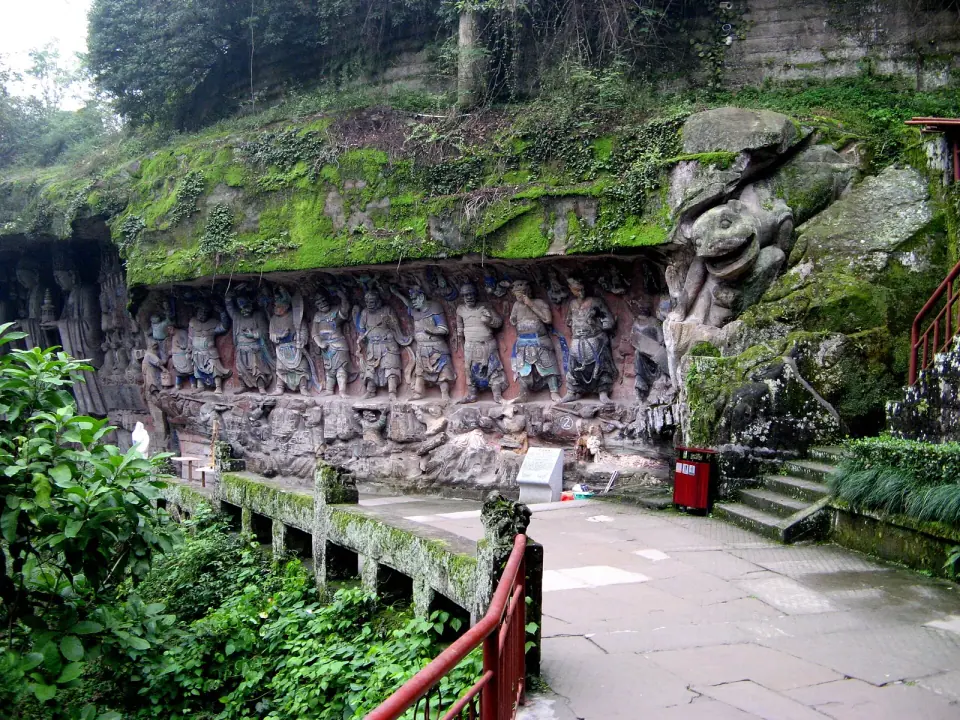 A long wall of Buddhist rock carvings beside a green hillside in Dazu
